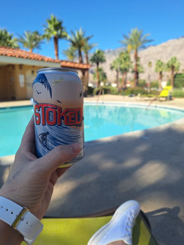 Hand holding a can of Stoked drink by a sunny pool with palm trees.