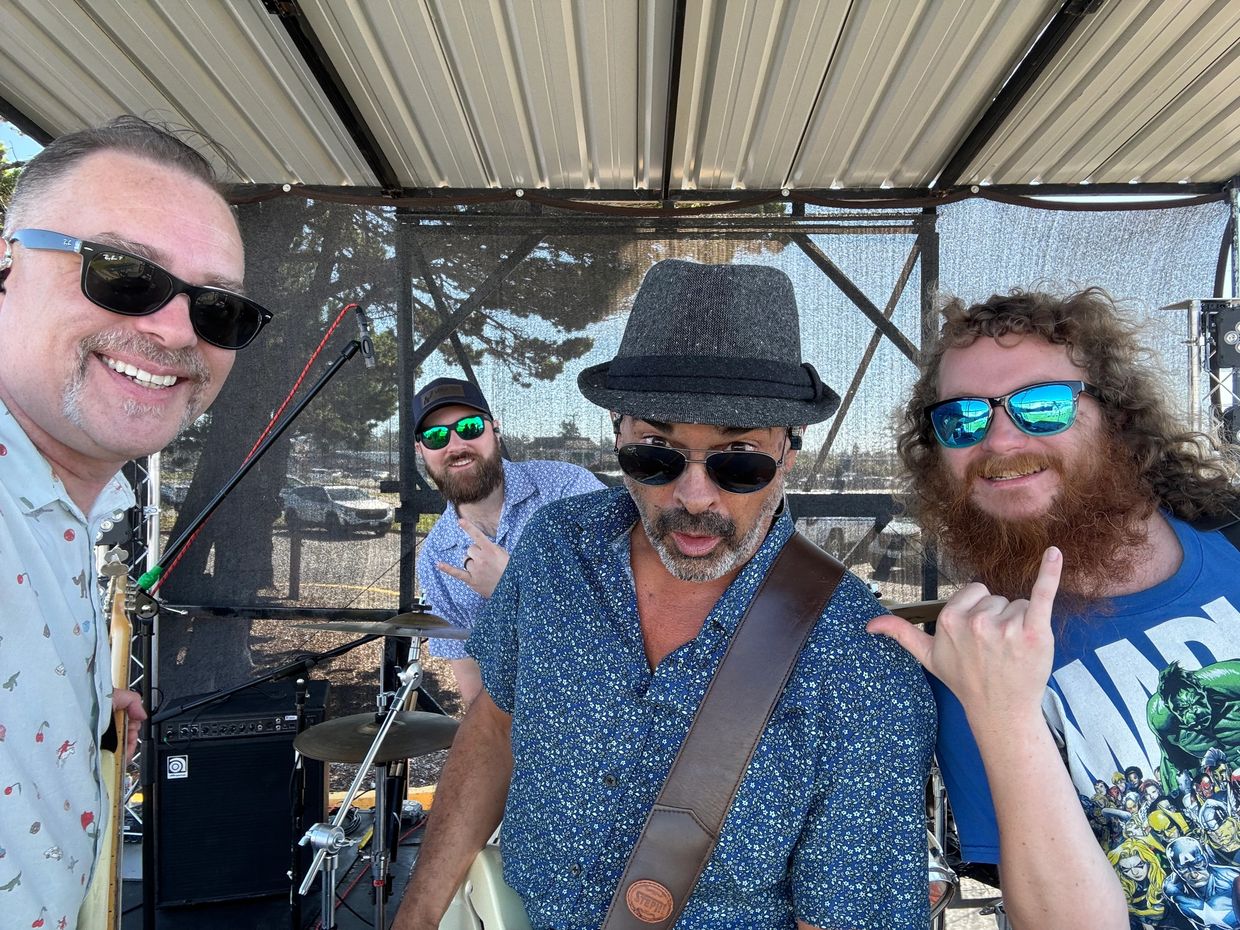 Four men in sunglasses posing under a canopy with musical instruments.