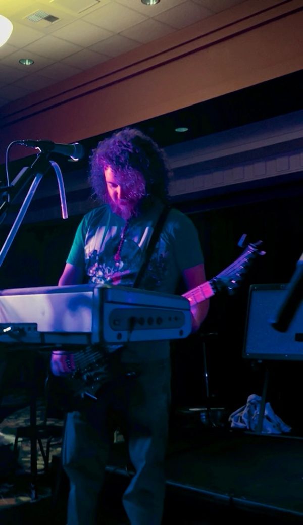 Musician with curly hair playing guitar and keyboard on stage under purple-blue lighting.