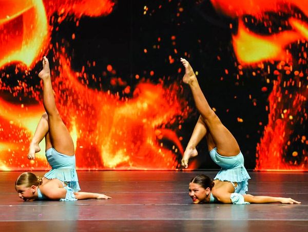 Two junior dancers in blue costumes doing chest stands onstage with a fire backdrop