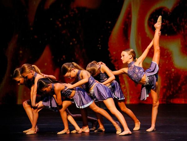 A young small group in muted purple costumes where one dancer is doing a needle