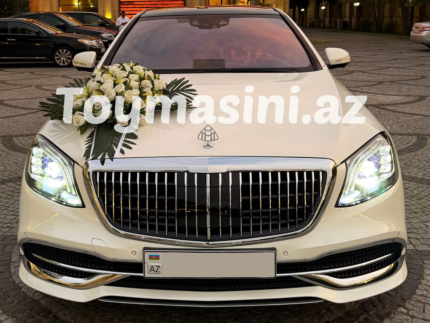 Elegant white Maybach with floral decoration on hood, headlights on.