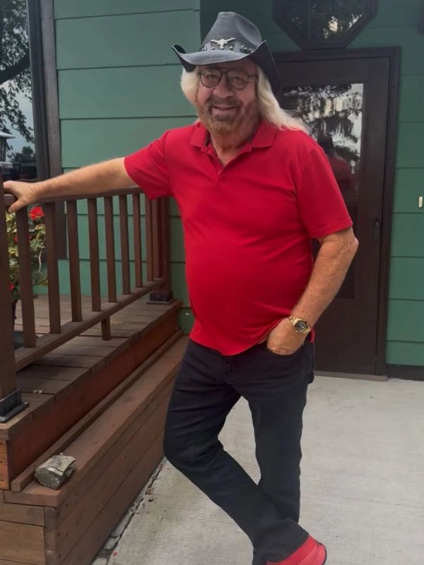 Man in red shirt and black cowboy hat leaning on wooden railing.
