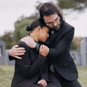A man and woman in black suits comforting each other at a cemetery.