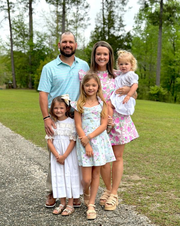 A smiling family of five posing outdoors on a green lawn with trees in the background.