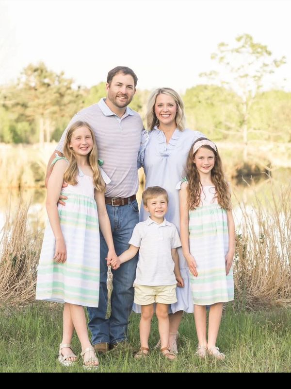 A smiling family of five posing outdoors near a pond on a sunny day.
