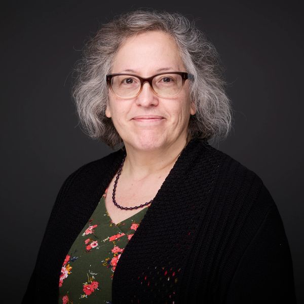 Smiling woman with gray hair and glasses wearing a floral dress and black cardigan.