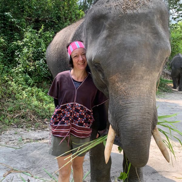 A smiling woman stands close to a large elephant eating grass.