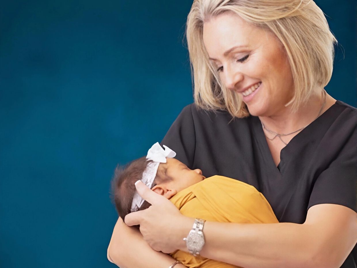 A smiling woman lovingly holds a swaddled baby against a deep blue background.