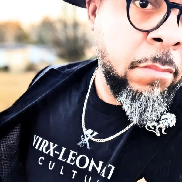 Man with glasses and a beard wearing a black shirt and jacket outdoors.