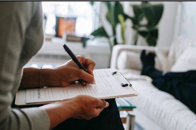 Person writing on a clipboard with someone lying on a couch.