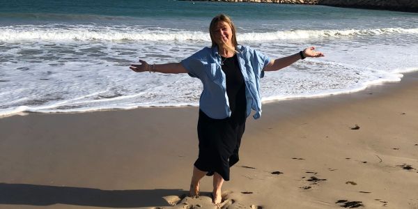 Woman joyfully standing barefoot on a sandy beach with waves in the background.