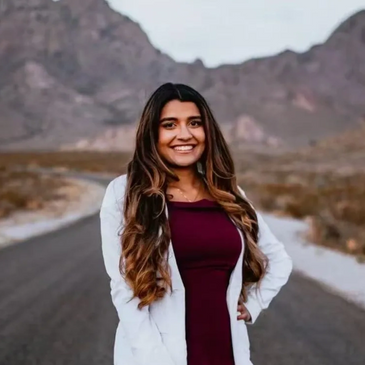 A woman stands smiling on a deserted road with mountains in the background.