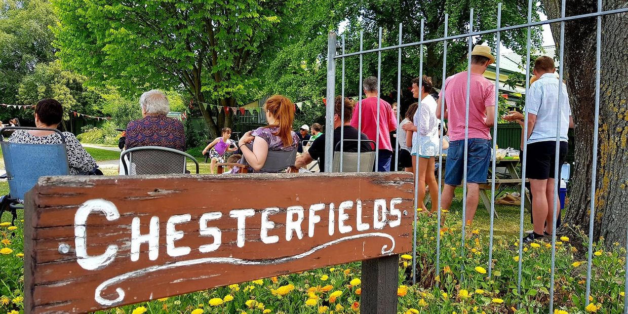 People gathered outdoors near a wooden Chesterfields sign in a garden setting.