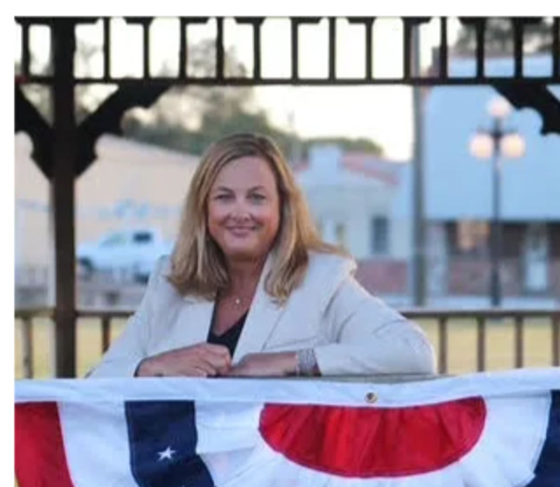 A woman in a white blazer smiling behind a decorated banner outdoors.