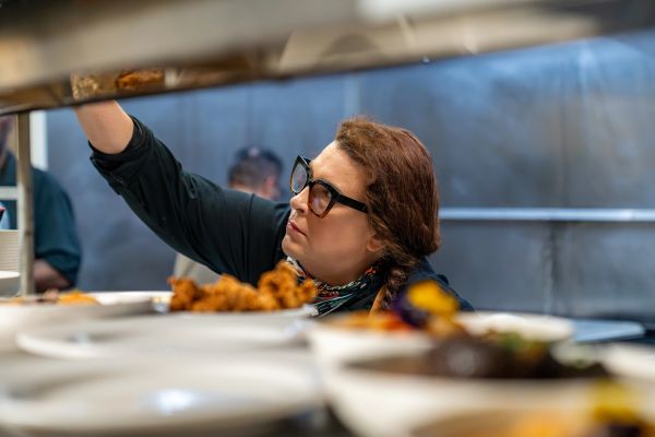 A chef wearing glasses reaches for something in a busy kitchen.