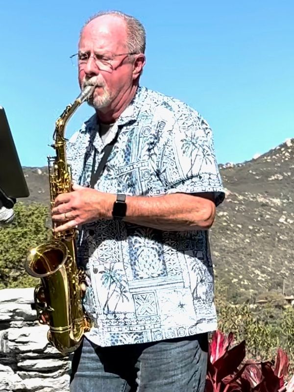 Man playing saxophone outdoors with mountains in the background.