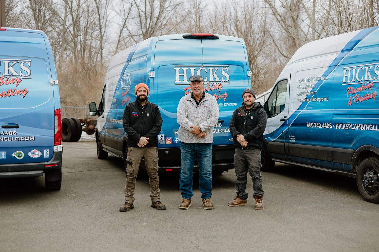 Three men standing in front of Hicks Plumbing &amp; Heating vans outdoors.