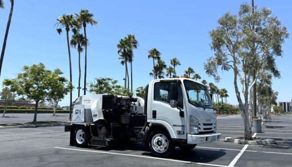 White street sweeper truck parked in an empty lot under palm trees.