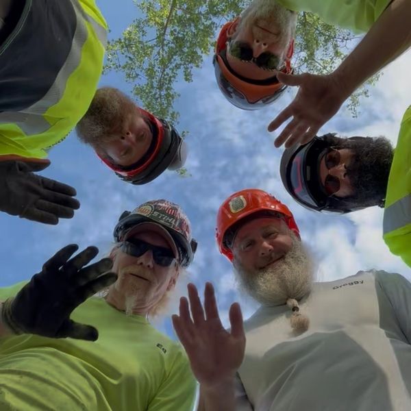 Five workers in helmets and safety gear wave at the camera outdoors.