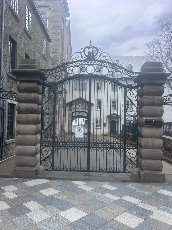 Ornate iron gate with stone pillars in front of historic building under cloudy sky.