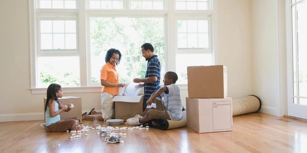 A family of four unpacking boxes together in a bright, empty living room with hardwood floors.