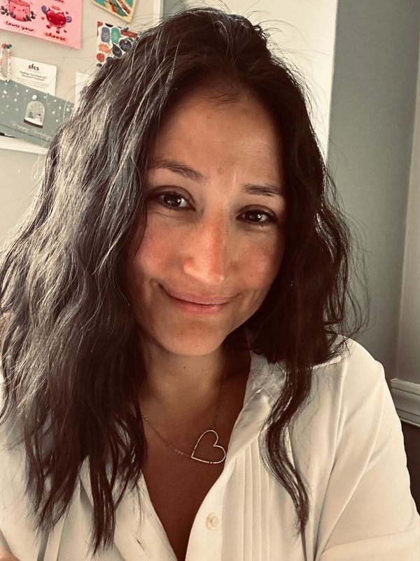 Smiling woman with wavy hair wearing a white blouse and heart necklace.