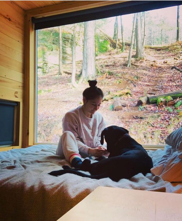 A woman sitting on a bed with a black dog by a large window overlooking the woods.