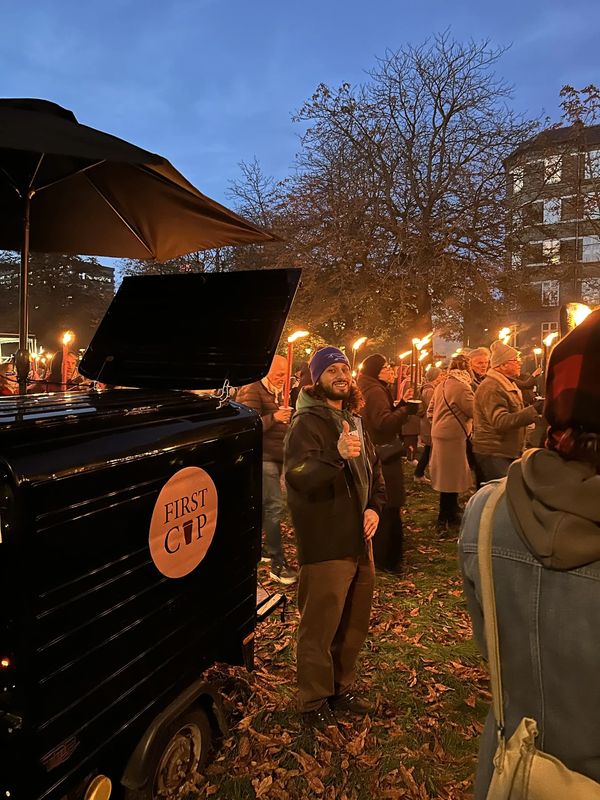 A man gives a thumbs up near a coffee cart at a torch-lit gathering in a park during dusk.
