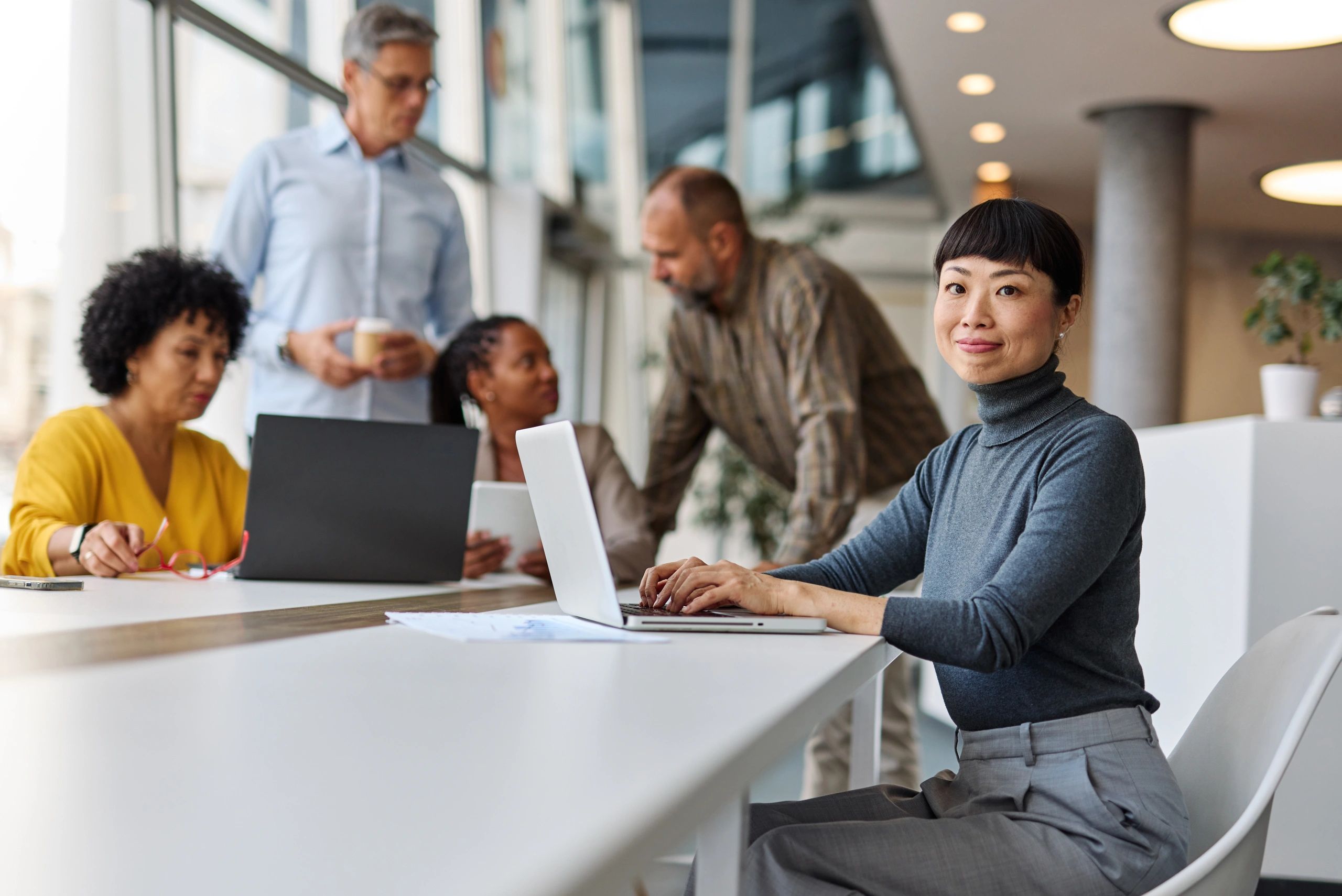 A confident woman working on a laptop in a modern office with colleagues collaborating in the background.