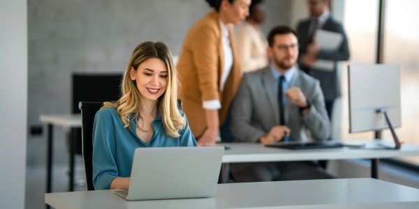Smiling woman working on a laptop in a modern office with colleagues.