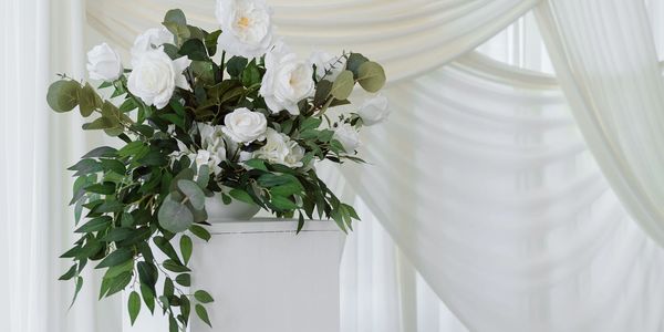 Elegant white floral arrangement on pedestal with soft draped curtains.