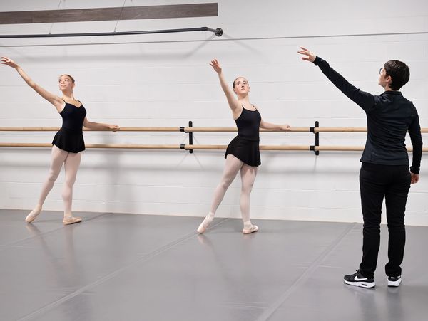 Two ballerinas practicing at the barre with an instructor guiding them.