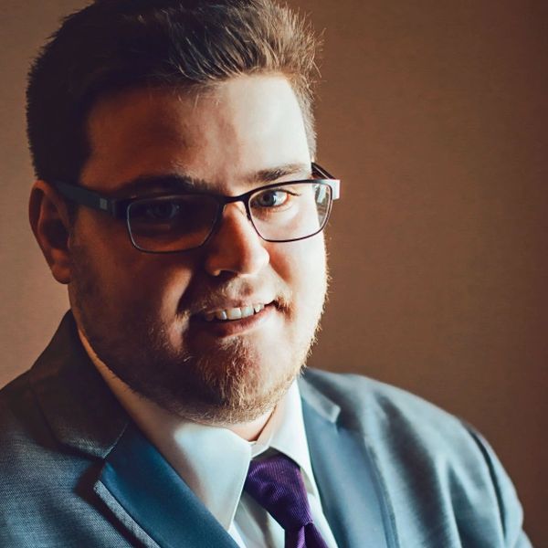Man wearing glasses and a suit smiles warmly in soft lighting.