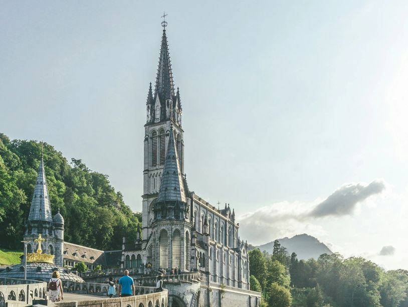 A grand Gothic-style church surrounded by lush greenery and mountains under a clear sky.