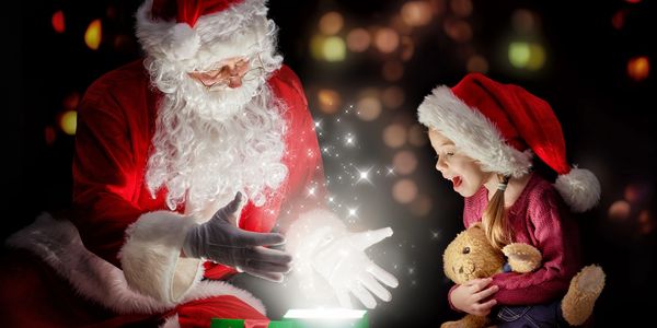 Santa and a little girl holding a teddy bear looking into a gift box that is glowing white.