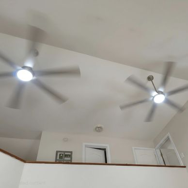 Two ceiling fans spinning with lights on in a room with white walls.