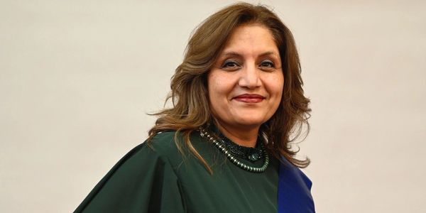 Woman in a dark green dress wearing a blue sash with gold fringe, smiling.