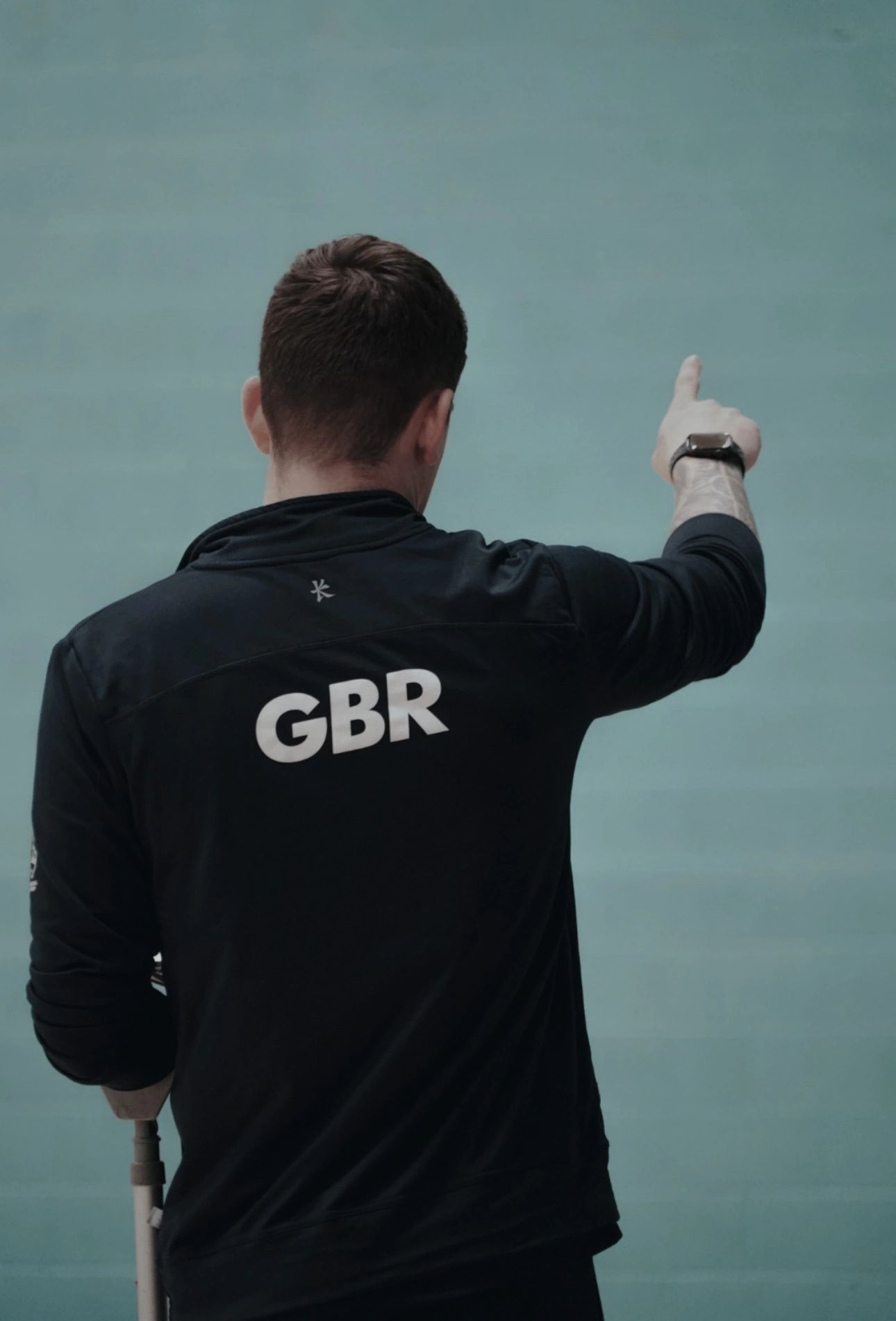 Man wearing GBR jacket points forward with a crutch in hand.