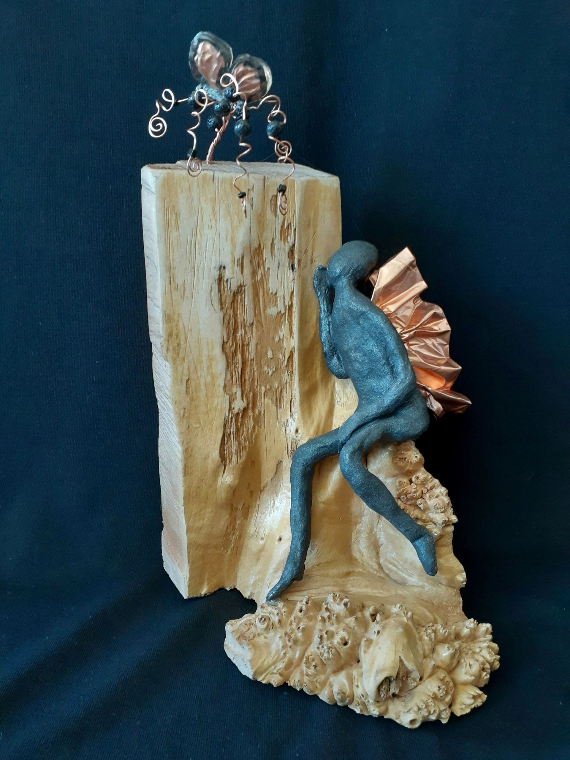 Abstract sculpture of a figure with butterfly wings sitting on wood.