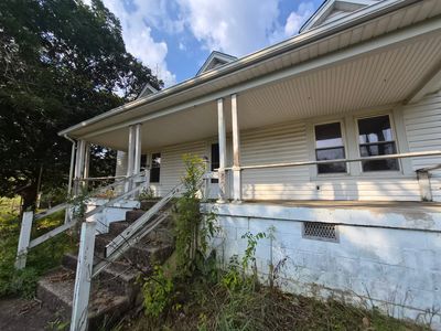 Old white house with a porch and overgrown plants.