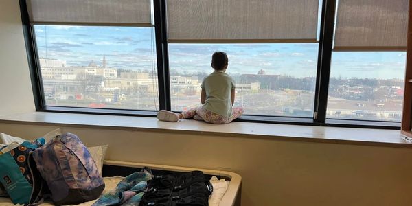 A child undergoing cancer treatment sits alone in her room, looking out of the window.