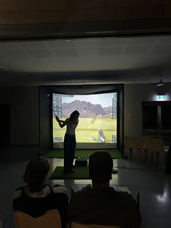 Person playing golf on an indoor golf simulator with two people watching.