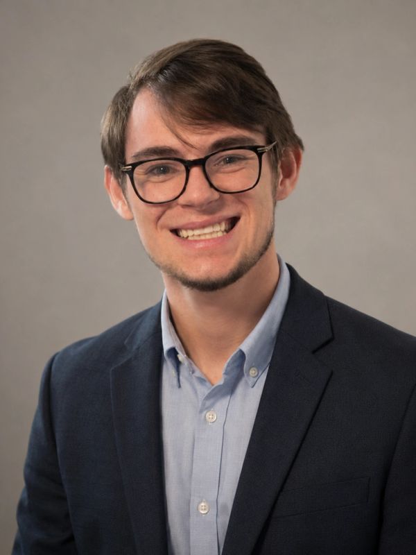 Young man in glasses and business attire smiling against a plain background.