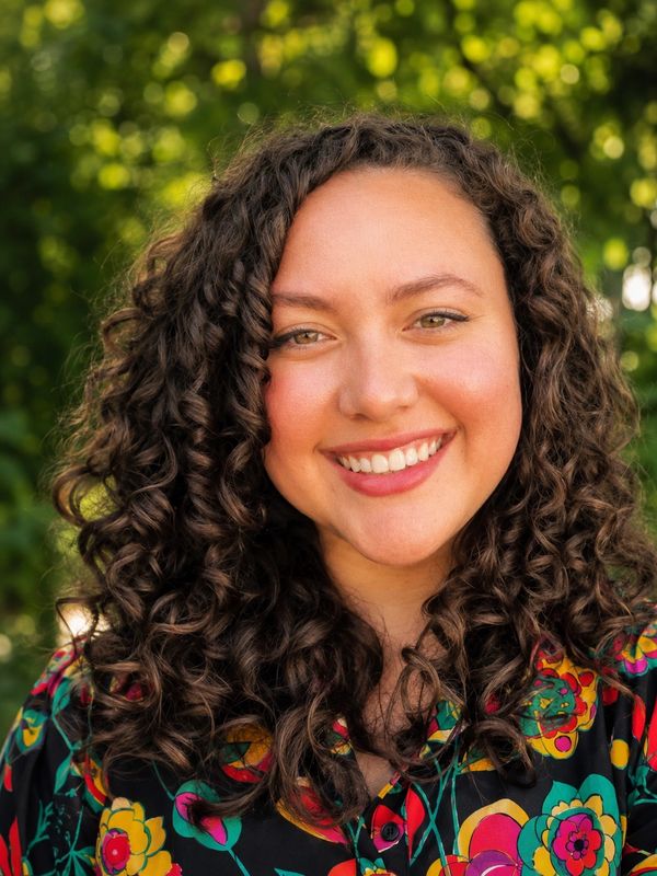 Smiling woman with curly hair and colorful floral shirt outdoors.