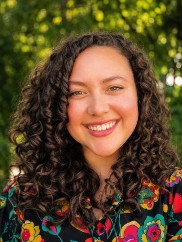 Smiling woman with curly hair and colorful floral shirt outdoors.