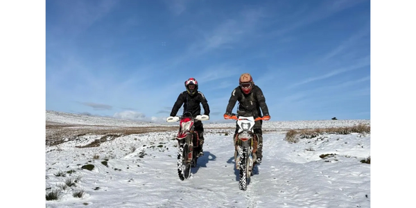 Two motorcyclists ride dirt bikes on a snowy landscape under a clear blue sky.
