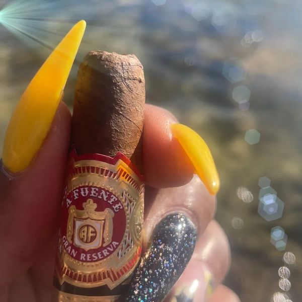 Hand holding an Arturo Fuente Gran Reserva cigar with vibrant yellow nails in sunlight.