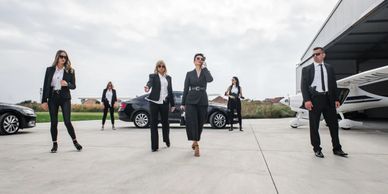 A team of six professionals in suits walking confidently on a tarmac near cars and a private jet.