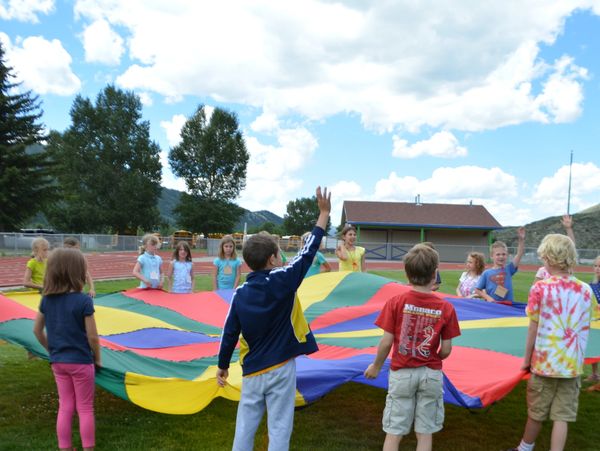 Children playing with a colorful parachute outdoors on a sunny day.